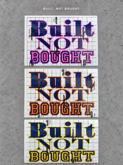 Three Built Not Bought vinyl decals with pink, orange and gold outlines and glossy blue lettering on grey concrete