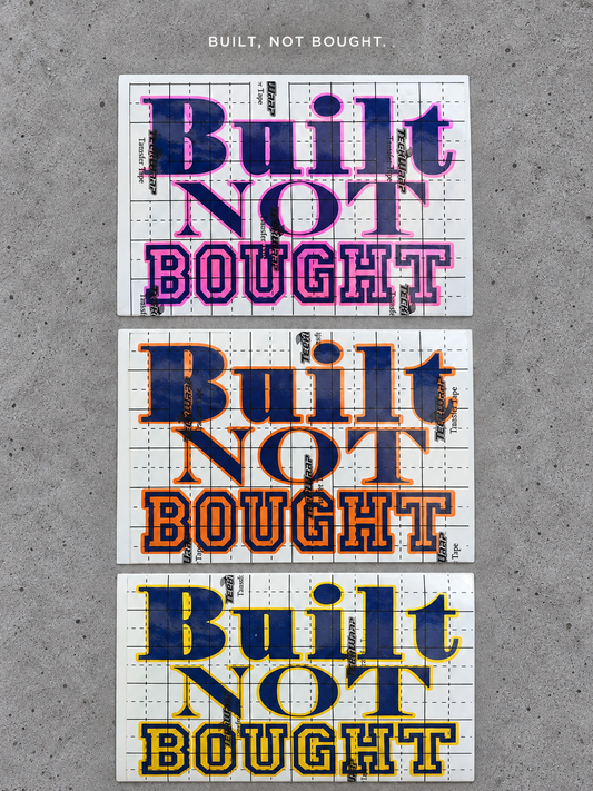 Three Built Not Bought vinyl decals with pink, orange and gold outlines and glossy blue lettering on grey concrete