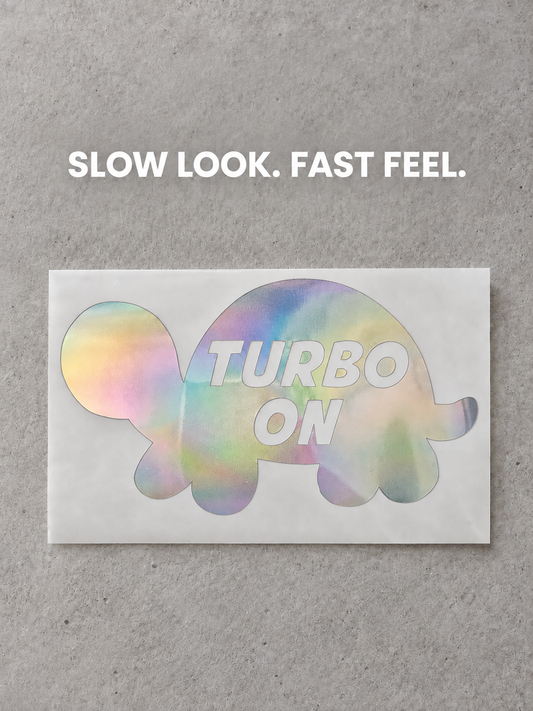 Turbo On turtle vinyl decal in soft pastel iridescent colour on grey concrete, with the white quote Slow Look. Fast Feel.