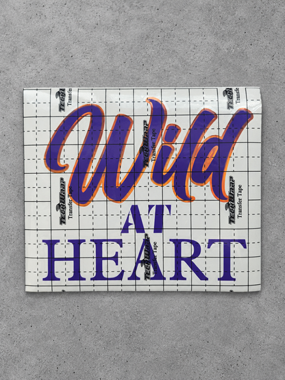 Wild at Heart vinyl decal in glossy purple with cayenne orange outline under transfer tape on grey concrete